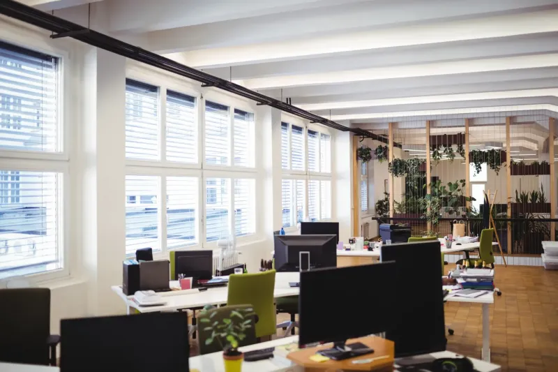 Großes, helles Büro mit mehreren Schreibtischen, Computermonitoren und grünen Bürostühlen vor großen Fenstern mit weißen Jalousien
