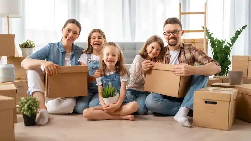 Familie mit zwei Erwachsenen und zwei Kindern sitzt auf dem Boden eines hellen Raums umgeben von Umzugskartons und Pflanzen